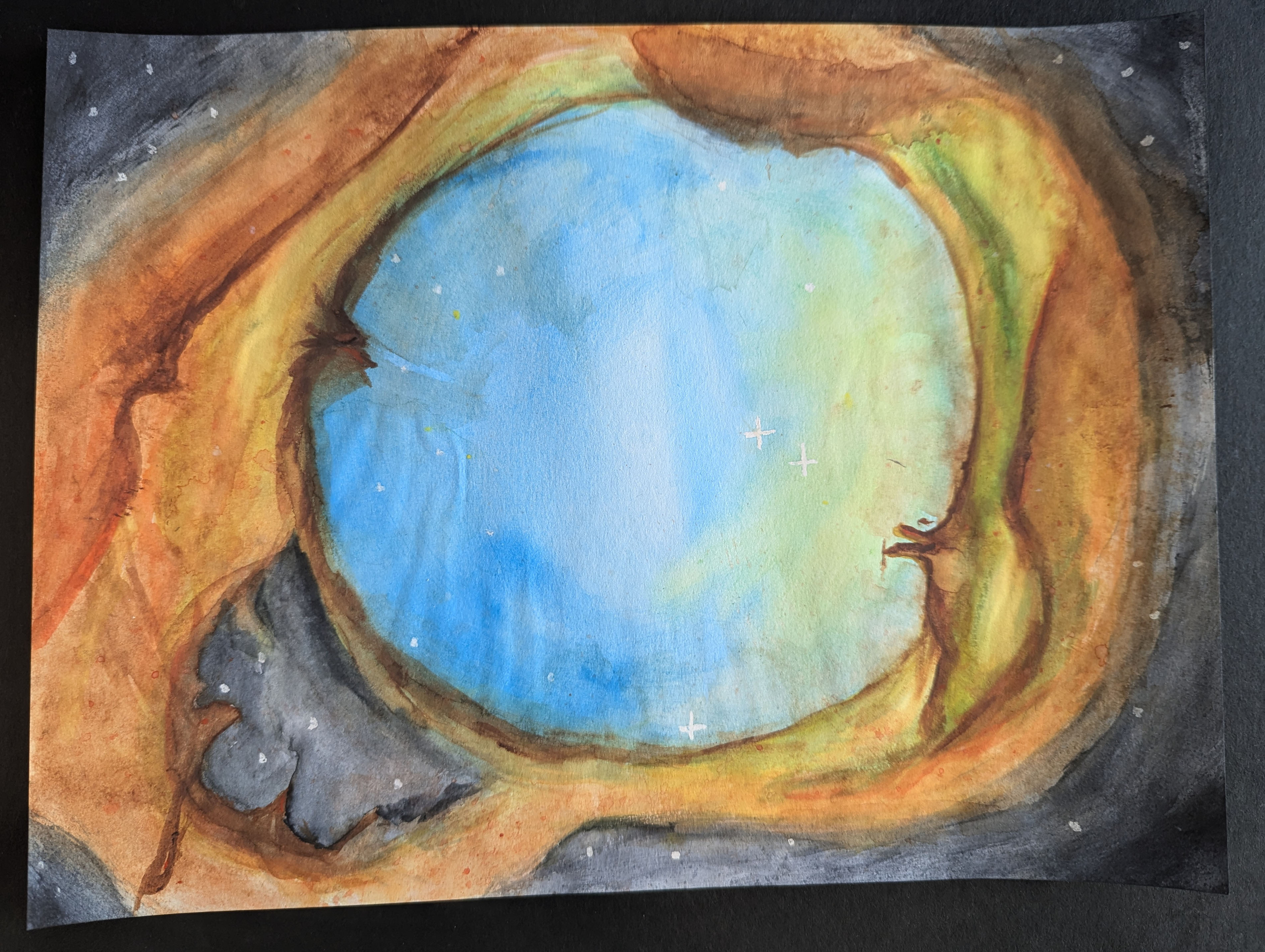 Watercolor painting of a star-forming nebula against a black tabletop. The nebula is a distorted oval that has a roughly circular blue and green center with a few stars, some with diffraction spikes. The immediate surrounding area is orange with some sections of green. In the lower left of the nebula is a gray cavernous area. The edges of the painting show the dark background of space with stars.