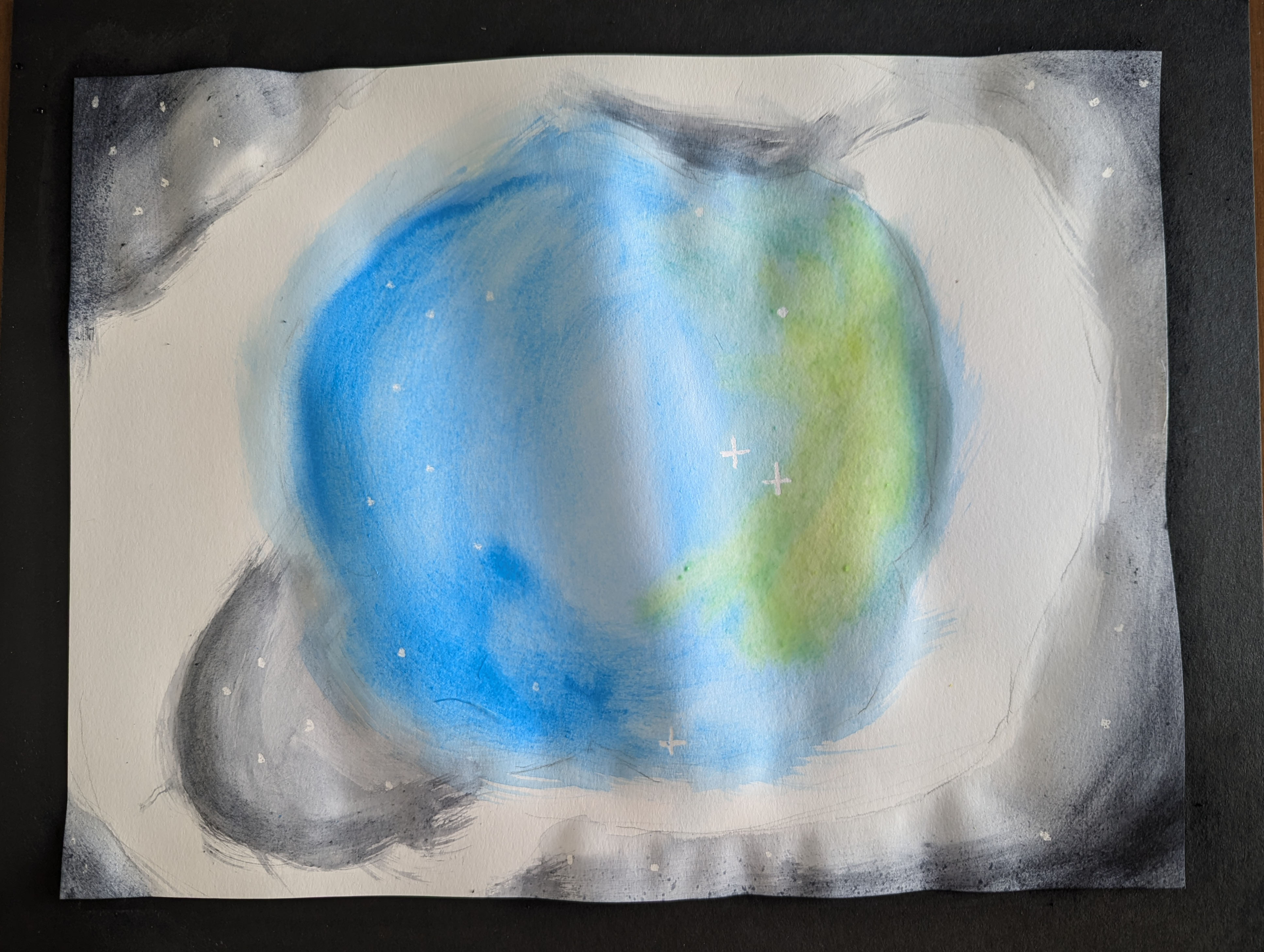 Photo of a white sheet of watercolor paper against a black tabletop. The edges of the paper have been painted dark gray, and there are stars. A large circle painted blue and green is in the center.