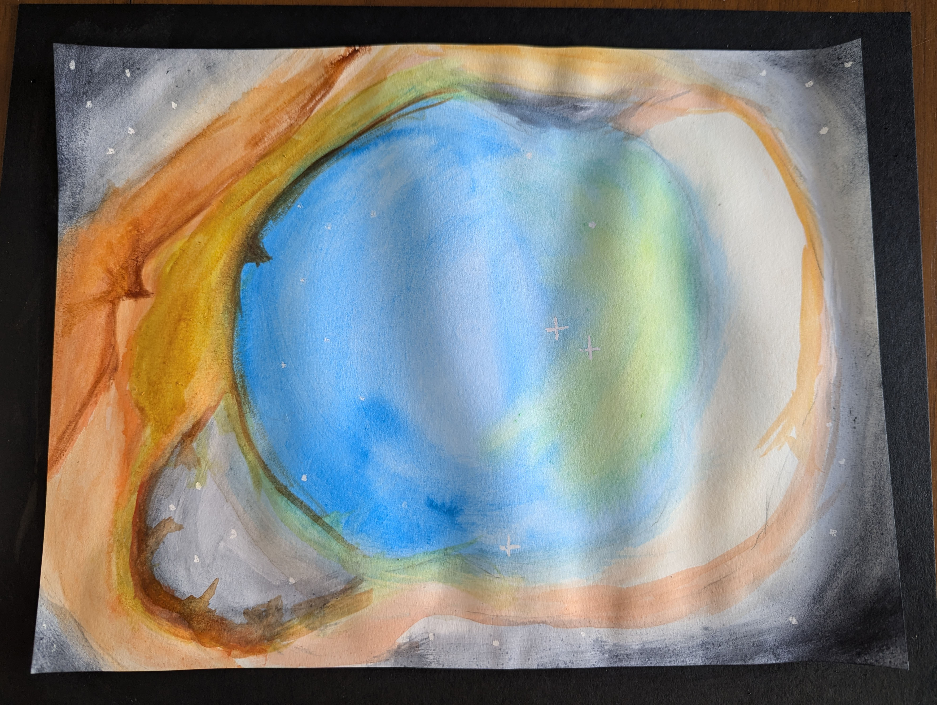 Photo of a watercolor painting against a black tabletop. The edges of the paper have been painted dark gray, and there are stars. A large circle painted blue and green is in the center. The immediate area surrounding the center is orange.