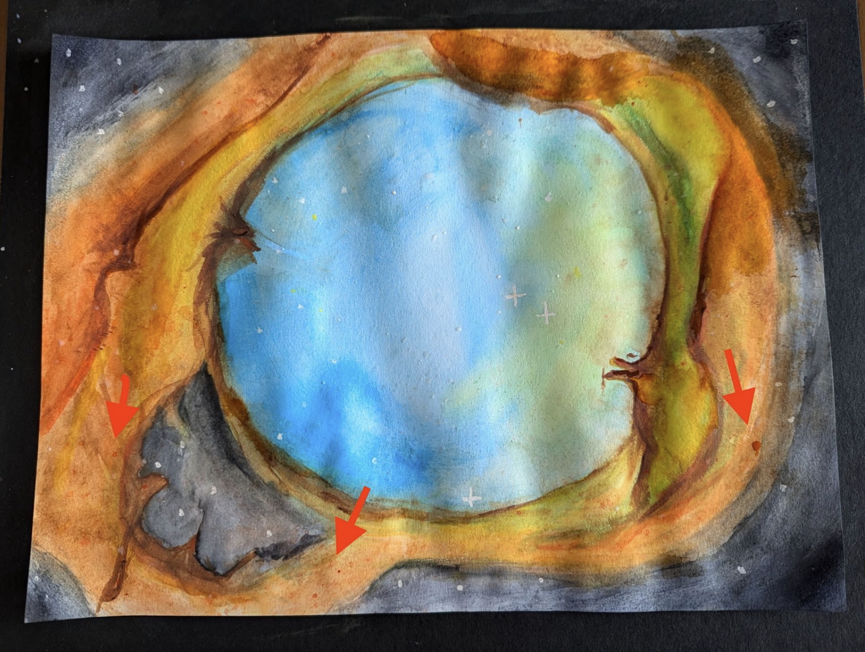 Photo of a watercolor painting against a black tabletop. The edges of the paper have been painted dark gray, and there are stars. A large circle painted blue and green is in the center. The immediate area surrounding the center is orange. Within the orange area are embedded stars that appear red.