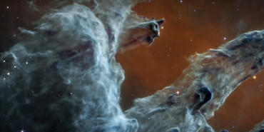 Semi-opaque layers of blue and gray gas and dust start at the bottom left and rise toward the top right. The background is deep red. A few red stars appear within the pillars, and some blue and white stars dot the scene.