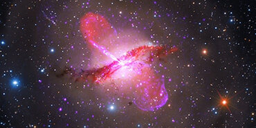 Against the dark background of space, a thin, wavy line of dark red material stretches diagonally across the center and extends from slightly lower left to slightly upper right. Purple and red bulbous plumes and lines are perpendicular to the dark red material and expand outward from the center toward the top left and bottom right. Hazy light brown concentric rings of light extend from the center, each ring becoming more transparent toward the image’s edges. Bright points of light in various sizes—some with diffraction spikes and halos—are densely scattered across this swath of space in various colors: purple, white, blue-white, yellow, and orange.