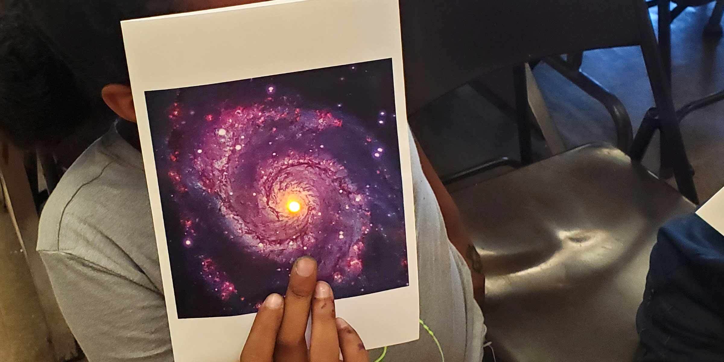 A Black child in a gray t-shirt holds a page in front of their face. The page shows a purple spiral galaxy rotating clockwise.