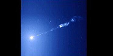 An image of M87’s jet observed by the Hubble Space Telescope whos a bright white circle at the bottom left corner, while a thin stream of material stretches diagonally across toward the upper right corner. The object lies against a blue background.