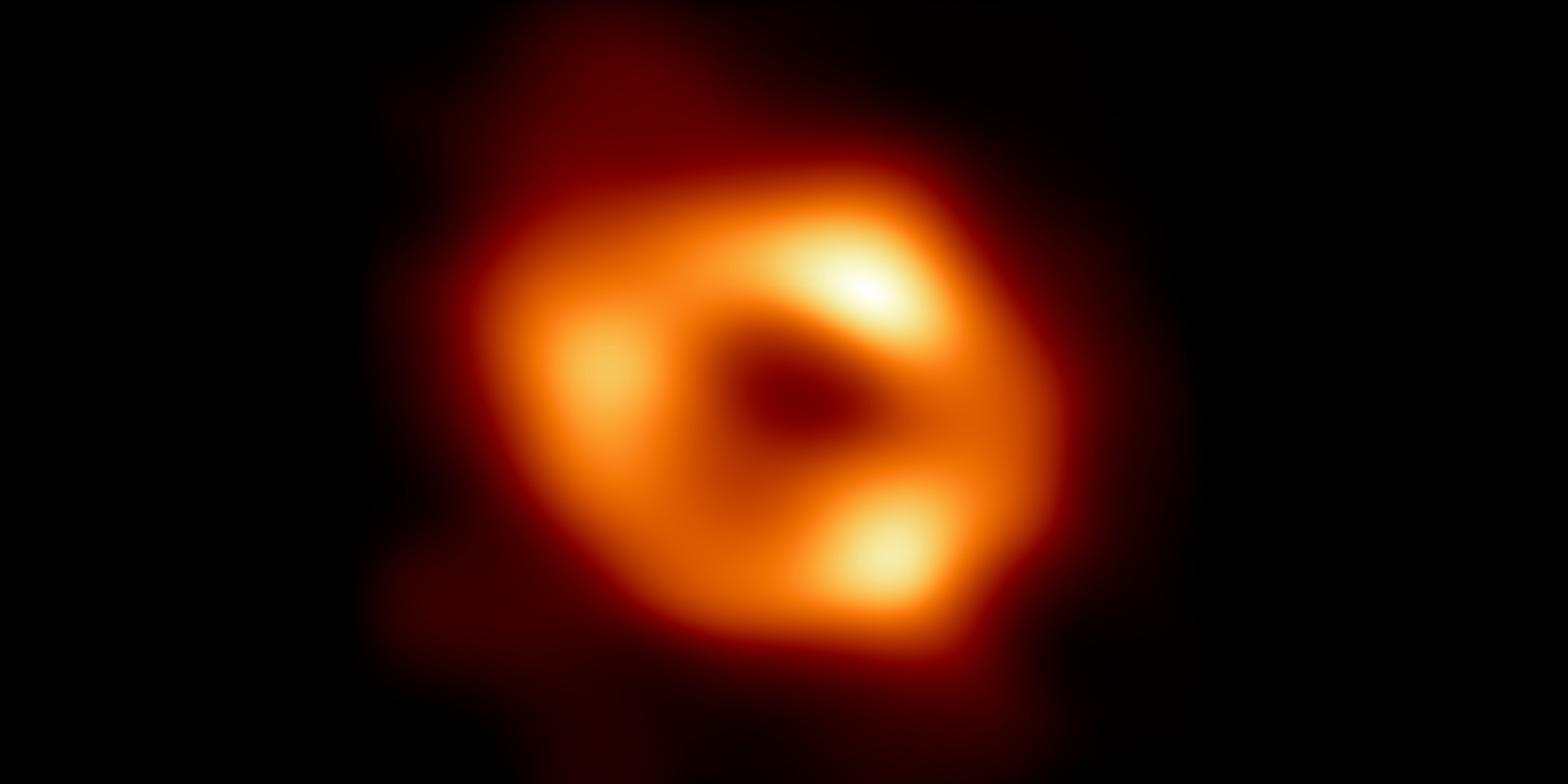 Event Horizon Telescope image of the black hole at the center of the Milky Way galaxy, Sagittarius A. It resembles a slightly oblong donut-shaped ring of glowing, warm dust. Within the ring, there are especially bright areas at the top, left, and right.
