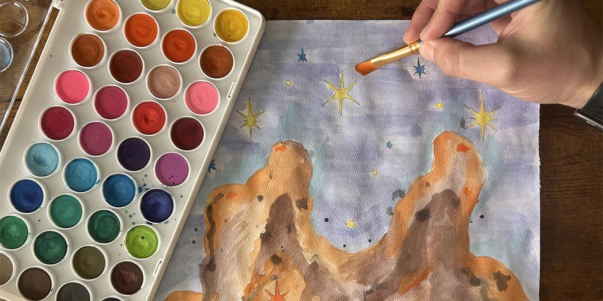 A person’s hand holds a paintbrush and is adding finishing touches to a watercolor painting of a star-forming nebula. The scene is viewed from above, with the paper placed on a wooden table. The nebula painting shows towering, reddish-brown pillar-like formations resembling those found in deep space images, surrounded by a soft, swirling purple and blue sky filled with stars. Bright yellow and orange star shapes, along with small scattered black and blue dots, depict stars scattered across the cosmic scene. On the left side of the image, a watercolor palette features over 30 vibrant colors, including shades of pink, orange, blue, green, and brown. A few colors show signs of recent use, with drops of paint on the palette and paper.