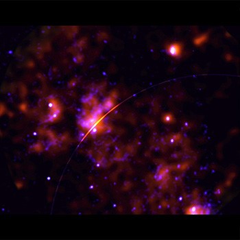 Center of the Milky Way galaxy as seen by the Chandra X-ray Observatory and NASA’s Imaging X-ray Polarimetry Explore. Dappled orange mist overlaps with cloudy indigo veins, and light purple specks. These patches of veiny mist are molecular clouds. A thin orange line diagonally stretches across part of the image’s center.