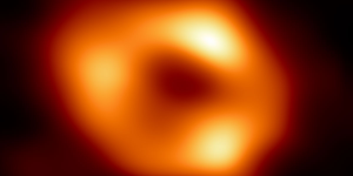 Event Horizon Telescope image of the black hole at the center of the Milky Way galaxy, Sagittarius A. It resembles a slightly oblong donut-shaped ring of orange glowing, warm dust. Within the ring, there are especially bright areas at the top, left, and right.