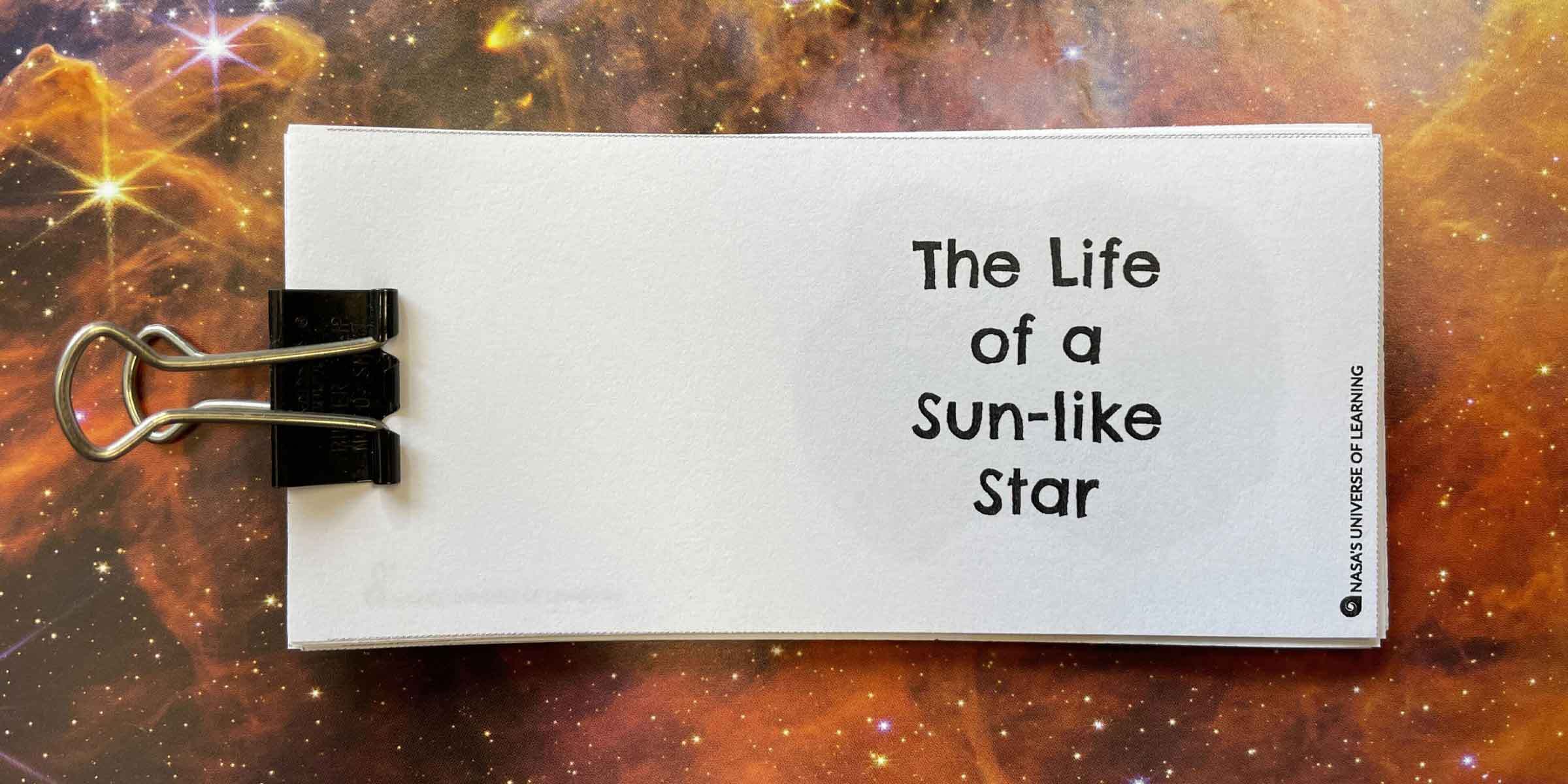 A printed horizontal series of white pages are clipped together at left by a black binder clip. The title on the first page, "The Life of a Sun-like Star" appears at right, with much smaller text running vertically, NASA's universe of Learning. The paper is set against a star field filled with stars and irregular layers of brown gas,