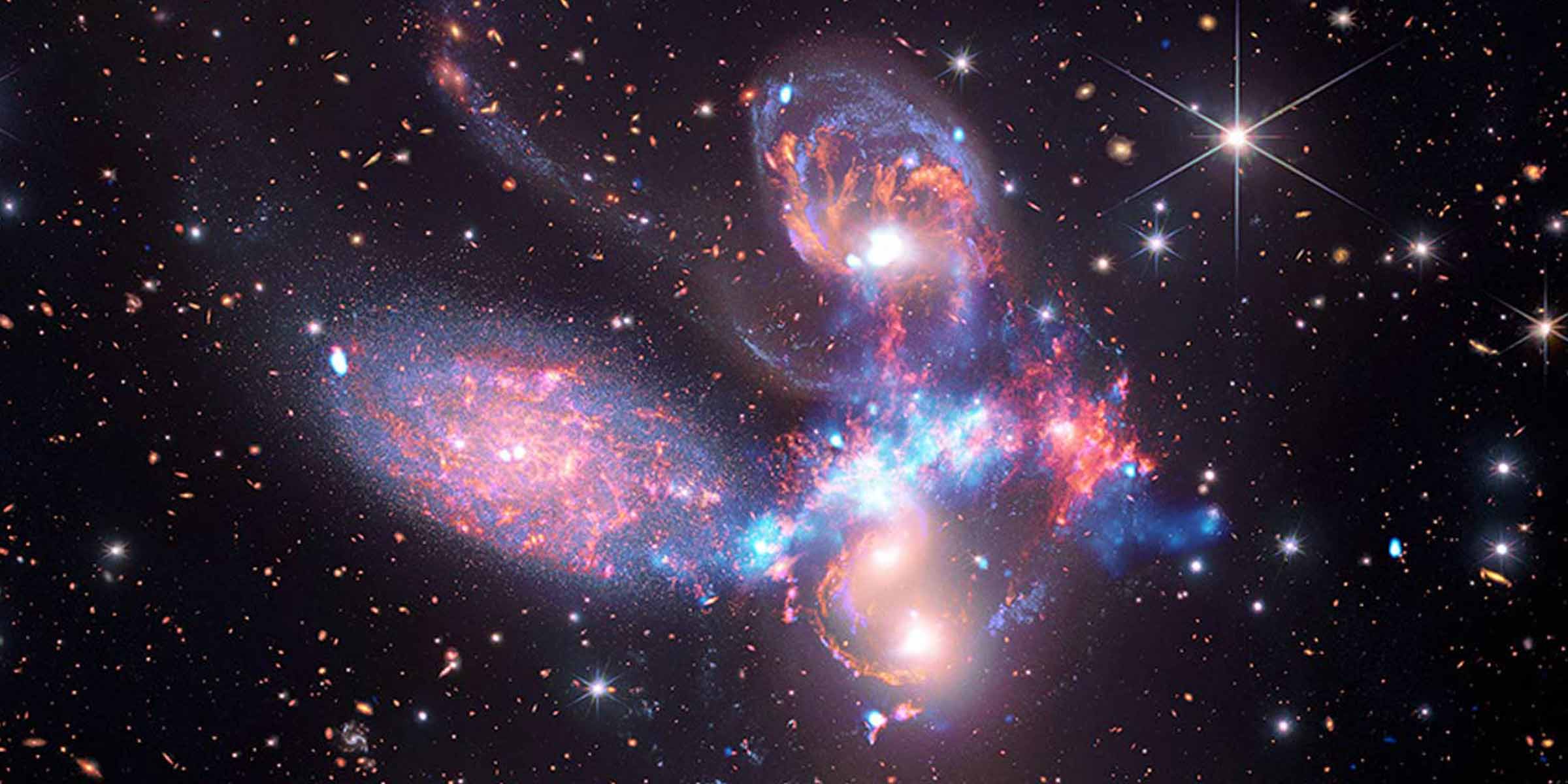 Four galaxies appear close to each other in the sky: two in the middle, one toward the top, and one to the upper left. The galaxies appear to be touching. The galaxies are large relative to the hundreds of much smaller (more distant) galaxies in the background. All four galaxies have bright white cores and have streaks of blue and red. Some of the galaxies are peppered with white dust-like specks. Each galaxy is unique in size, shape, structure, and exact coloring. Scattered across the image, in front of the galaxies, are a number of foreground stars with diffraction spikes: bright white points, each with eight bright lines radiating out from the center.