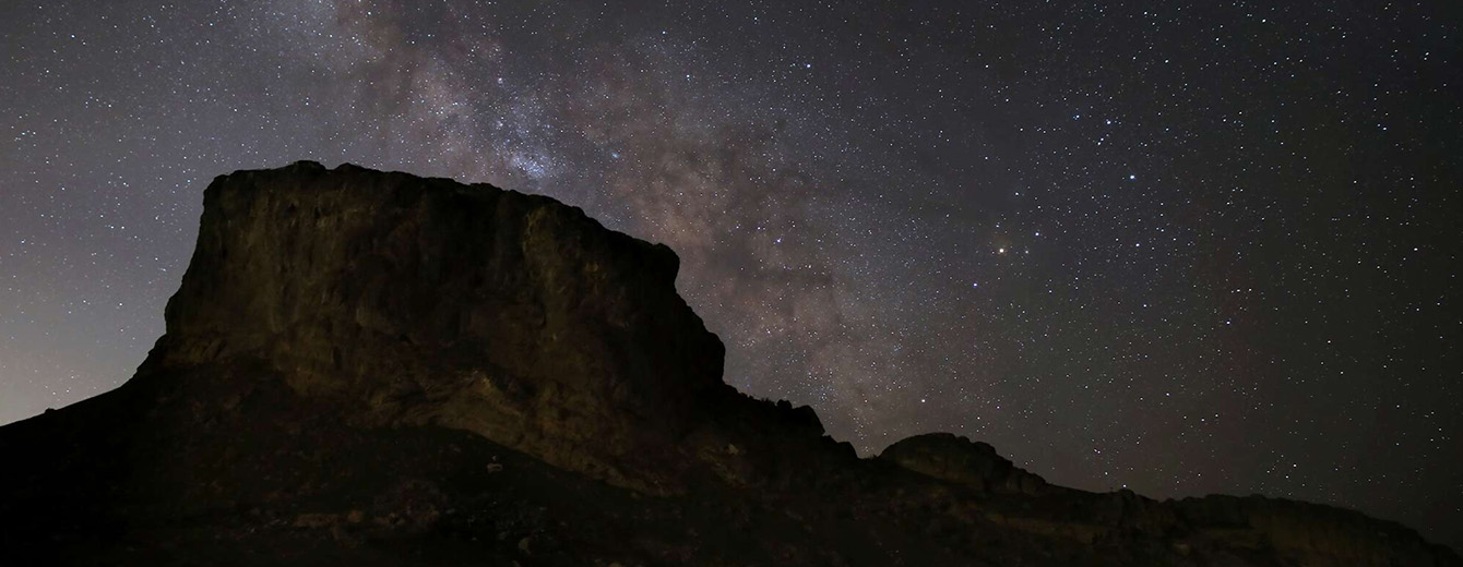 A photograph shows a desert butte in front of a night sky glittering with stars. The band of the Milky Way crosses the sky diagonally, from top left to middle bottom. It looks like a glowing cloud filled with dust and stars above the desert landscape.