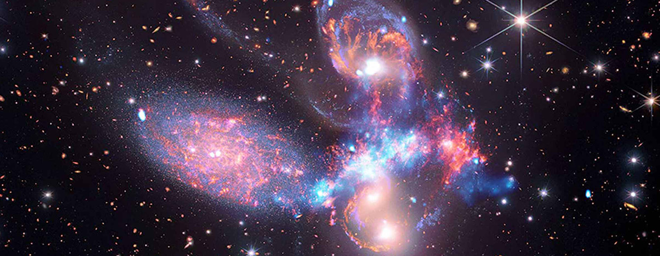 A glowing horizontal line sweeps across Stephan’s Quintet, a compact galaxy group. The five galaxies appear close to each other in the sky: two in the middle, one toward the top, one to the upper left, and one toward the bottom. Four of the five appear to be touching. The one toward the bottom is somewhat separated. The galaxies are large relative to the hundreds of much smaller (more distant) galaxies in the background. All five galaxies have bright white cores. The four galaxies toward the top of the image have streaks of blue and red. Some of the galaxies are peppered with white dust-like specks. Each galaxy is unique in size, shape, structure, and exact coloring. Scattered across the image, in front of the galaxies, are a number of foreground stars with diffraction spikes: bright white points, each with eight bright lines radiating out from the center.