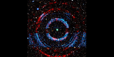 V404 Cygni. Blotchy blue and red material form ring-like structures around a blue-green dot in the center. In the background are many points of light scattered across a black swath of space, varying in size, color, and detail.