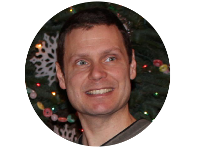 A close up headshot of a white male with short brown hair smiling. A garland of snowflakes and beads appears in the background.