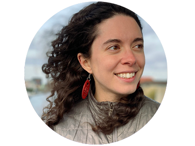 A white woman with curly brown hair and brown eyes, wearing a grey coat and red earrings, looking to the side.