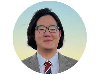 A man wearing black glasses, wearing a tweed suit, smiles at the camera in front of a sunset.