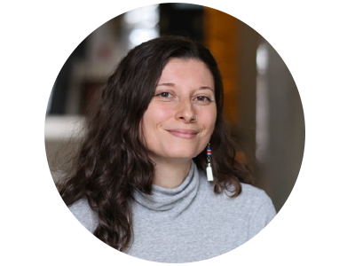 Headshot of a women with dark hair and a closed mouth smile wearing a grey high neck sweater and white earrings that dangle