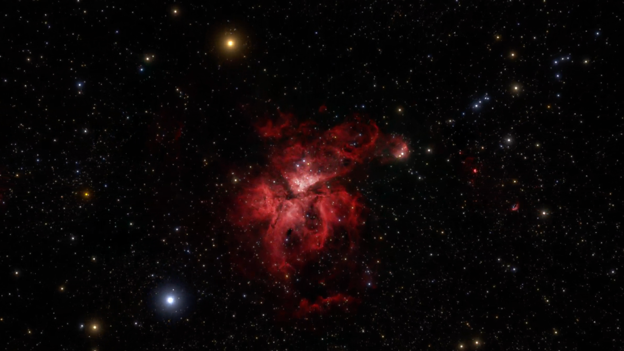 At the center is a loose splotch of red, the Carina Nebula, set against the black background of space. Distant stars and galaxies are speckled across the scene. The very center of the Carina Nebula is brightest, almost white, with larger stars represented as dots. The gas and dust that make up the nebula take the loose form of flower petals, like an iris. The top “petal” appears in the rough shape of a lowercase V and becomes more diffuse and darker red the farther away the material is from the core. Off to its right is a small, loose red circle with several bright white stars within it. The lower petal of the nebula is also deep red, appearing more like a triangle with one peak centered at the bottom. That edge peters out into deeper red and becomes more translucent the lower it goes. Below that, a short horizontal line of deep semi-transparent red material appears. There is a prominent orange orb center-left at the top, and a blue one toward the bottom a little farther left.